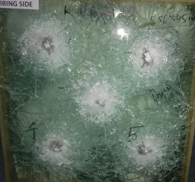 The firing side of a bullet resistant panel after a trial: five numbered impact craters, each ringed by a star of radial cracking, the pane otherwise whole.