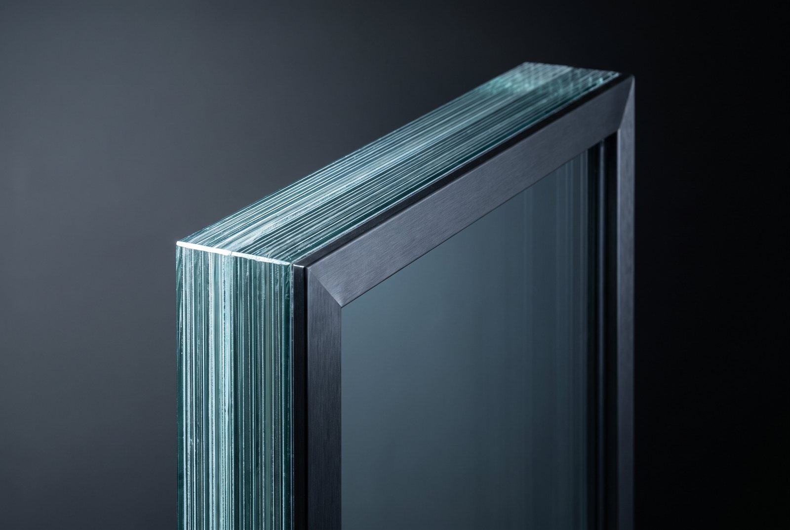 The corner of a thick multi-ply laminated glass panel held in a dark steel frame, the exposed edge showing the stack of glass plies and interlayers.