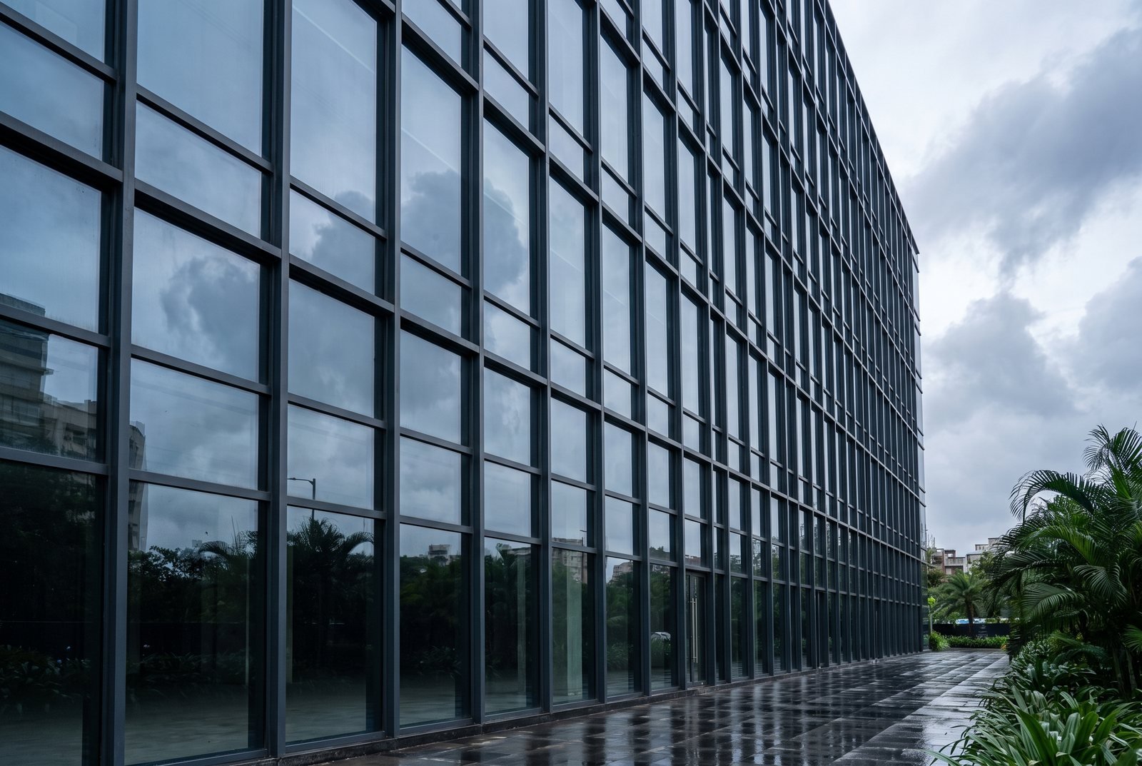 A multi-storey curtain wall facade in a slim dark steel grid seen at a raking angle from ground level, regular glazed bays running the full height of the elevation with an overcast sky reflected across them.