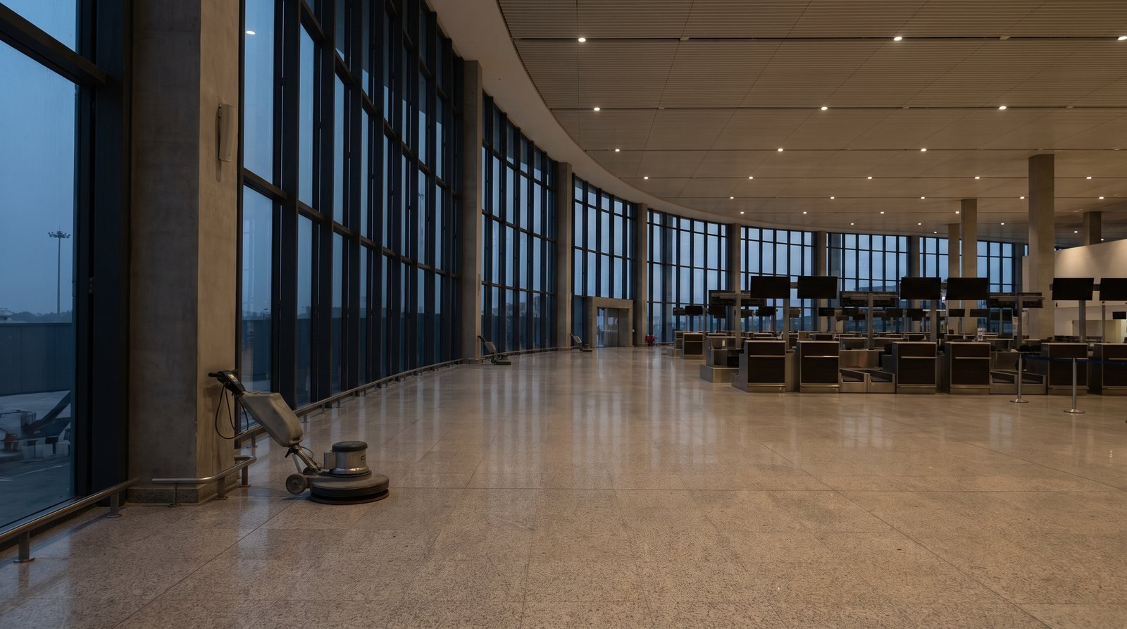 A curved glazed concourse wall in a slim dark frame, check-in desks along the far side and the apron visible through the glass.