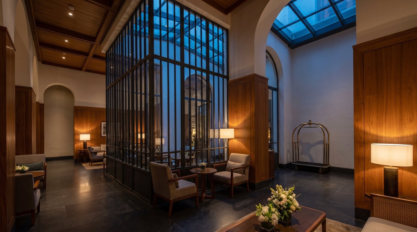A hotel lobby with a full-height steel-framed glazed screen dividing the seating area, timber panelling and a luggage trolley beyond it.